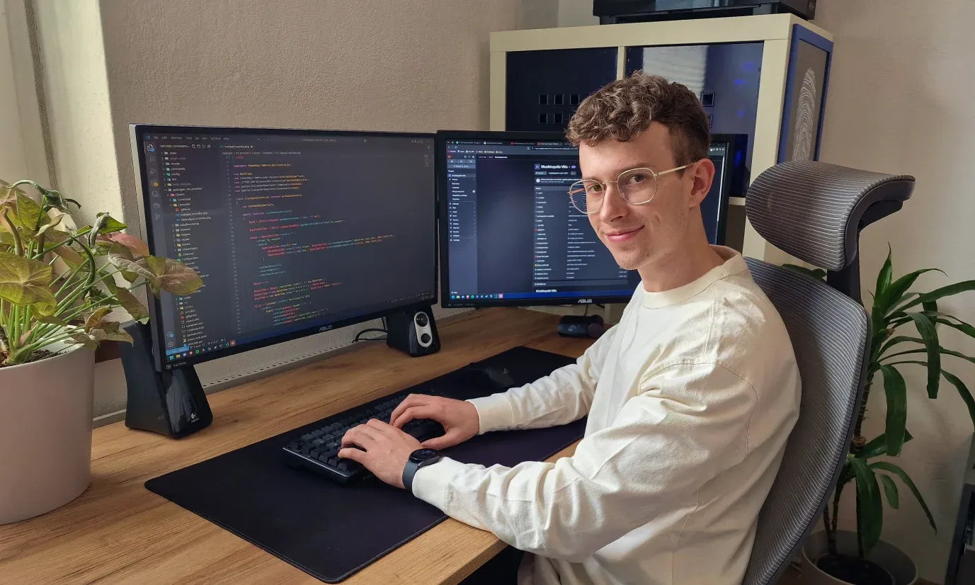 Simon Mayr an seinem Schreibtisch in Innsbruck, an zwei Bildschirmen mit TYPO3-Code und einer Kundenwebsite, umgeben von Pflanzen.